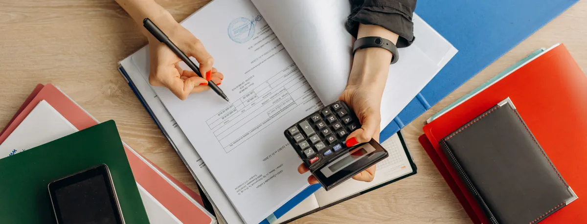 Hands going through invoices with a pen and a calculator on a desk of coloured folders (sample photo)