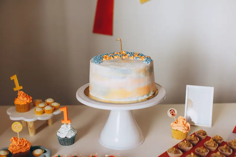 A pastel birthday cake on a white stand with cupcakes around it